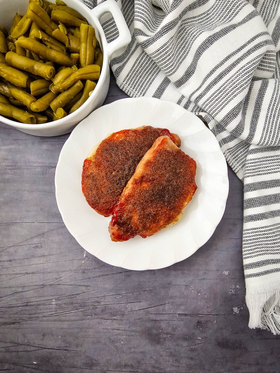 Looking down at a dinner plate with two sweet and savory pork chops next to a white bowl filled with green beans.