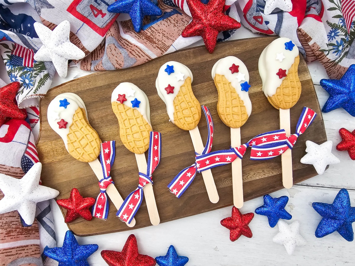 An overhead view of several patriotic nutter butter cookie pops neatly arranged on a wooden cutting board. The bright white coating and red and blue star sprinkles contrast beautifully with the dark wood grain and surrounding glittery star decorations.