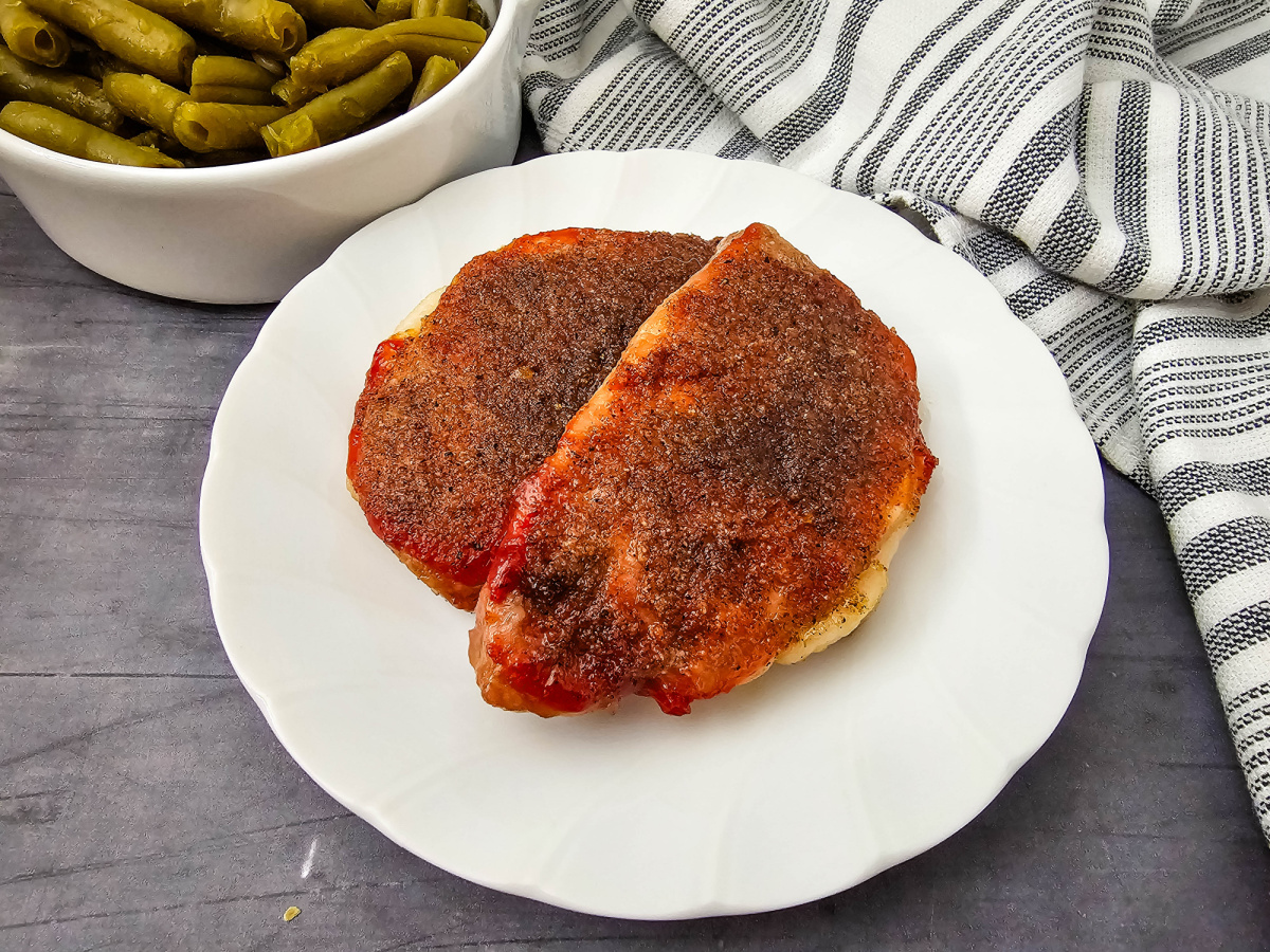 A close-up, side-angle view of two thick baked pork chops coated in a rich, caramelized brown sugar and ketchup glaze, served on a white plate with a bowl of green beans behind it.