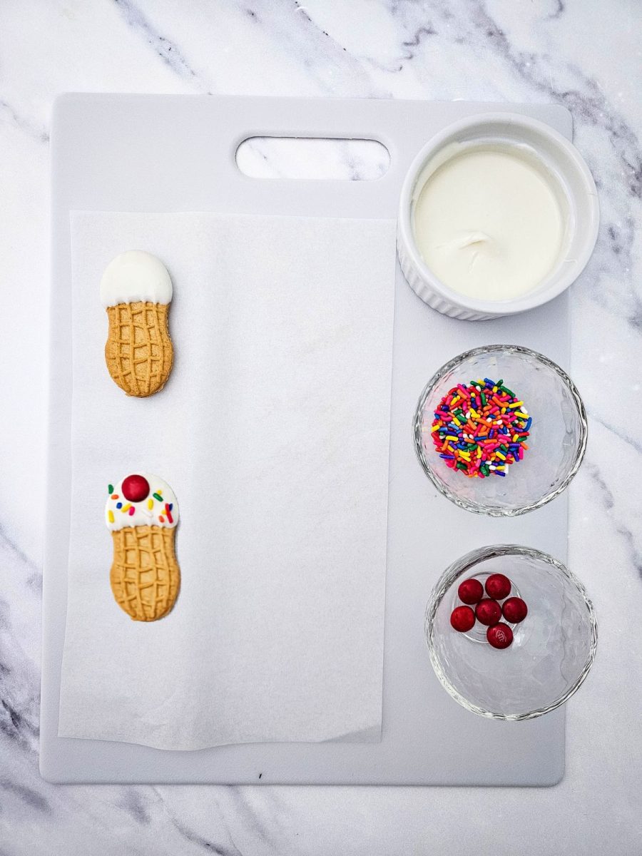 A step-by-step recipe process view showing a white cutting board lined with parchment paper, featuring one peanut butter cookie dipped in white melting wafers, one fully decorated cookie, a bowl of melted white chocolate, a bowl of sprinkles, and a bowl of red candies.