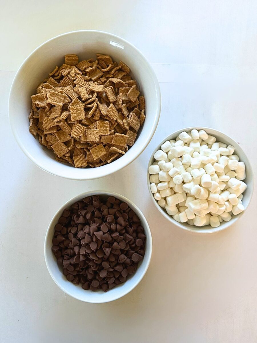 Three simple ingredients for making S'mores Snack Mix are arranged in bowls: Golden Grahams cereal, milk chocolate chips, and mini marshmallows.