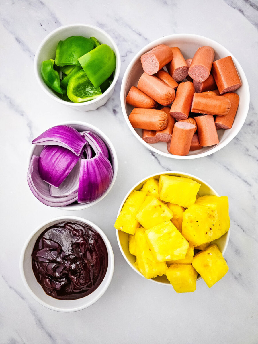 Small bowls hold sliced beef hot dogs, green bell peppers, red onion wedges, pineapple chunks, and barbecue sauce before making the kabobs. The ingredients are arranged on a marble countertop.