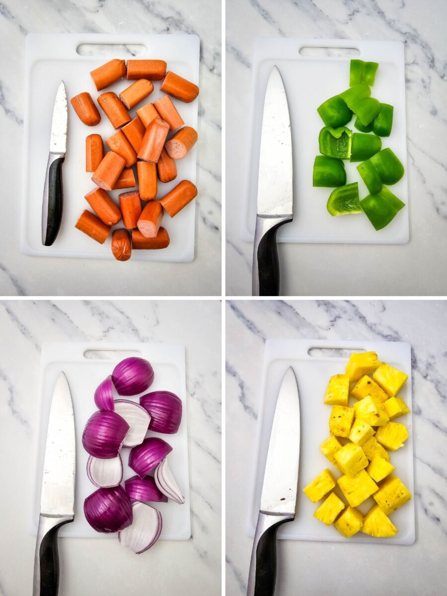 Four cutting boards display chopped beef hot dogs, green bell peppers, red onion wedges, and pineapple chunks before assembling the kabobs. A chef's knife rests beside each ingredient on a marble countertop.