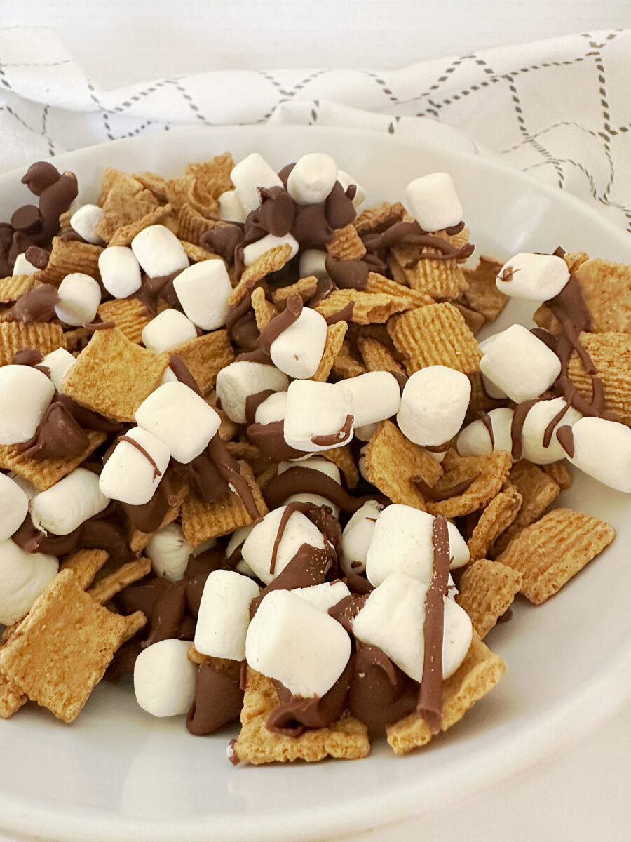 A close-up of S'mores Snack Mix in a white bowl shows Golden Grahams cereal coated with milk chocolate and mini marshmallows. 