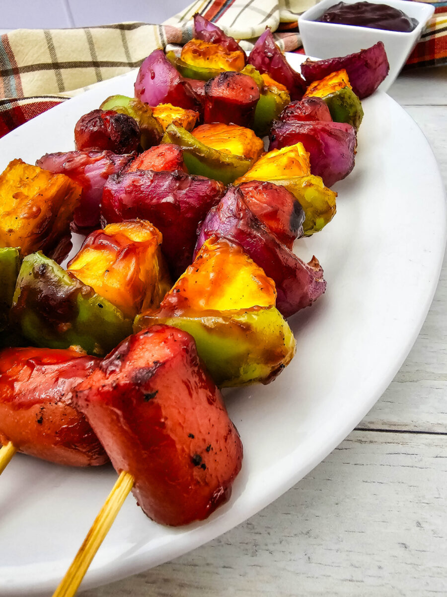 A close-up of grilled hot dog kabobs shows caramelized pineapple, charred hot dogs, green peppers, and red onion on a white serving platter. A small bowl of barbecue sauce and a plaid kitchen towel sit in the background.
