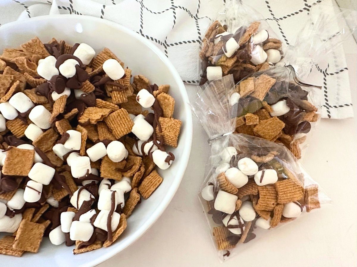 A bowl of S'mores Snack Mix is displayed next to two individual treat bags filled with the sweet cereal snack.