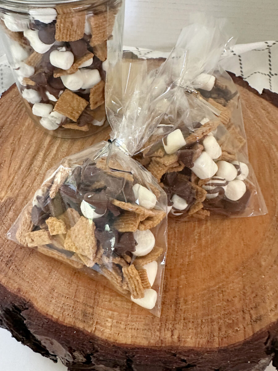 A glass jar filled with S'mores Snack Mix sits behind two individually packaged snack bags on a rustic wood slice. 