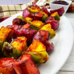 Three grilled hot dog kabobs are arranged diagonally on a large white platter with caramelized pineapple, green peppers, and red onion. A bowl of barbecue sauce and a plaid towel complete the tabletop setting.