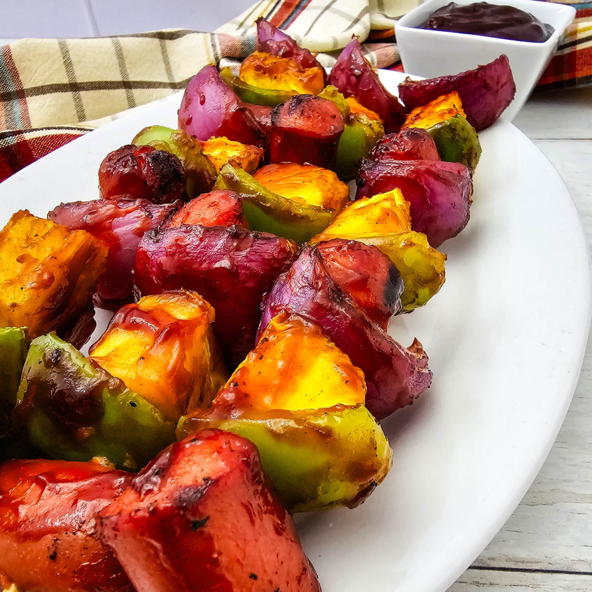 Three grilled hot dog kabobs are arranged diagonally on a large white platter with caramelized pineapple, green peppers, and red onion. A bowl of barbecue sauce and a plaid towel complete the tabletop setting.