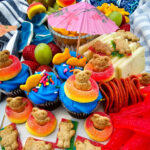 Pool party charcuterie board with cupcakes, fruit, cheese, crackers, Swedish fish candy, and goldfish crackers arranged on a summer snack board.