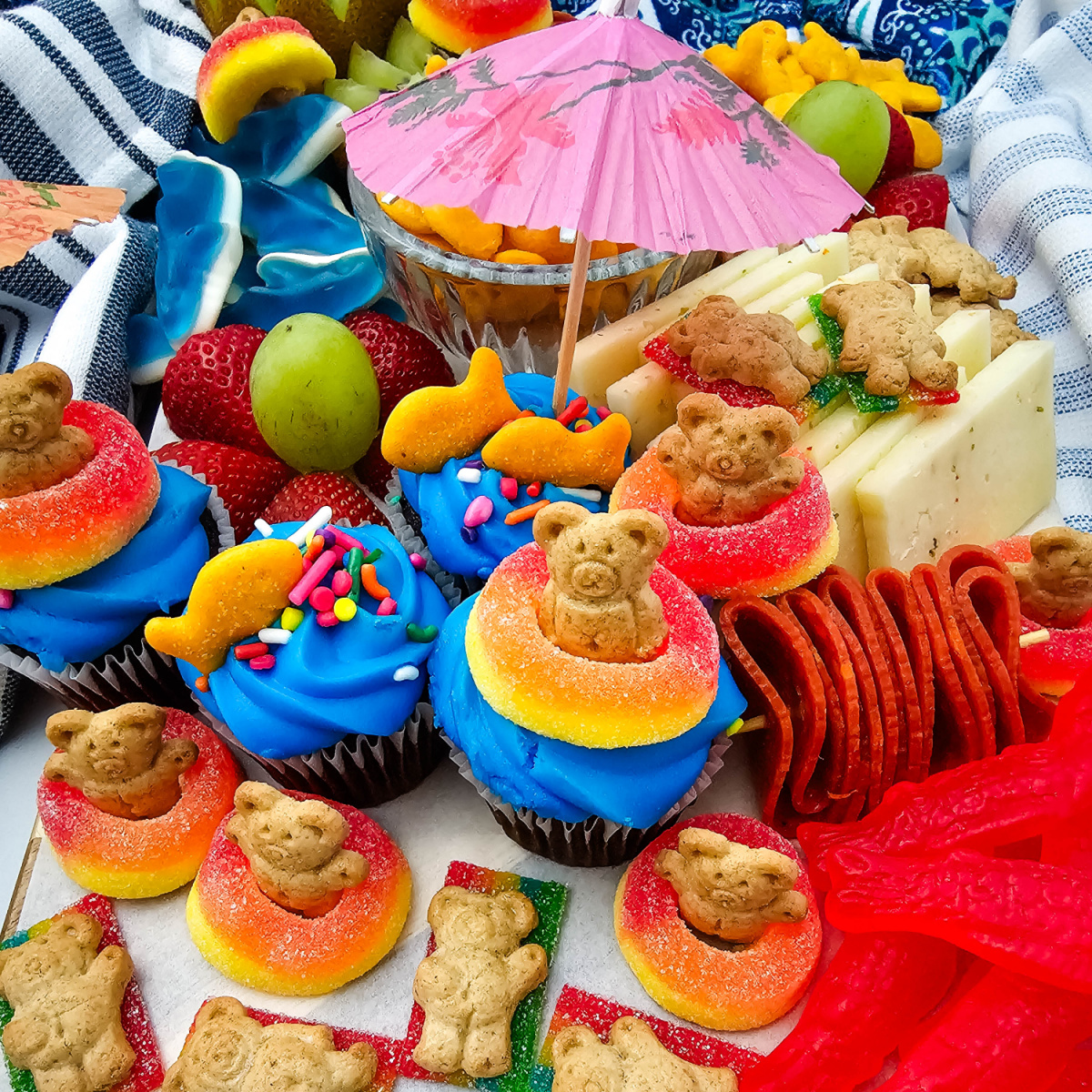 Pool party charcuterie board with cupcakes, fruit, cheese, crackers, Swedish fish candy, and goldfish crackers arranged on a summer snack board.