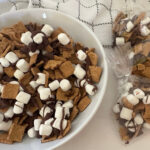 A bowl of S'mores Snack Mix sits beside individually packaged treat bags filled with the cereal snack mix. This easy sweet snack combines Golden Grahams cereal, milk chocolate, and mini marshmallows for a fun treat.