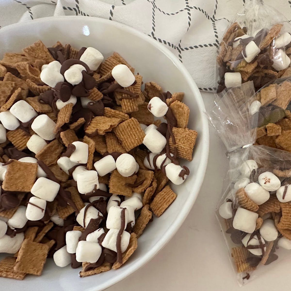 A bowl of S'mores Snack Mix sits beside individually packaged treat bags filled with the cereal snack mix. This easy sweet snack combines Golden Grahams cereal, milk chocolate, and mini marshmallows for a fun treat.
