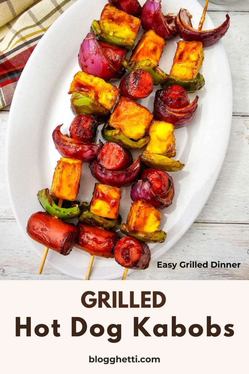 Pinterest graphic of grilled hot dog kabobs on a white serving platter with the text "Easy BBQ Dinner." The skewers are topped with grilled pineapple, peppers, and red onion beside a bowl of barbecue sauce.