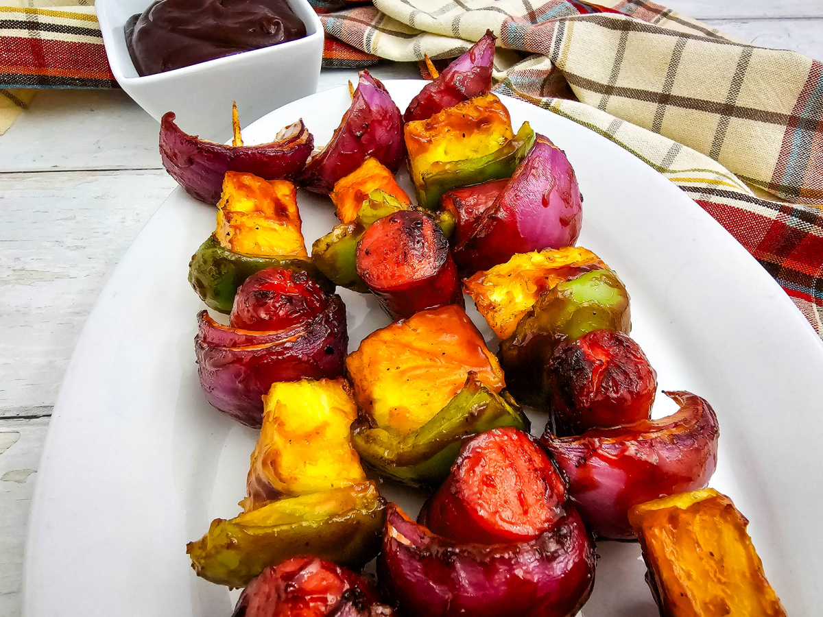 Three grilled hot dog kabobs rest on a white oval platter with charred pineapple, green peppers, and red onion. A bowl of barbecue sauce sits behind the platter next to a plaid kitchen towel.