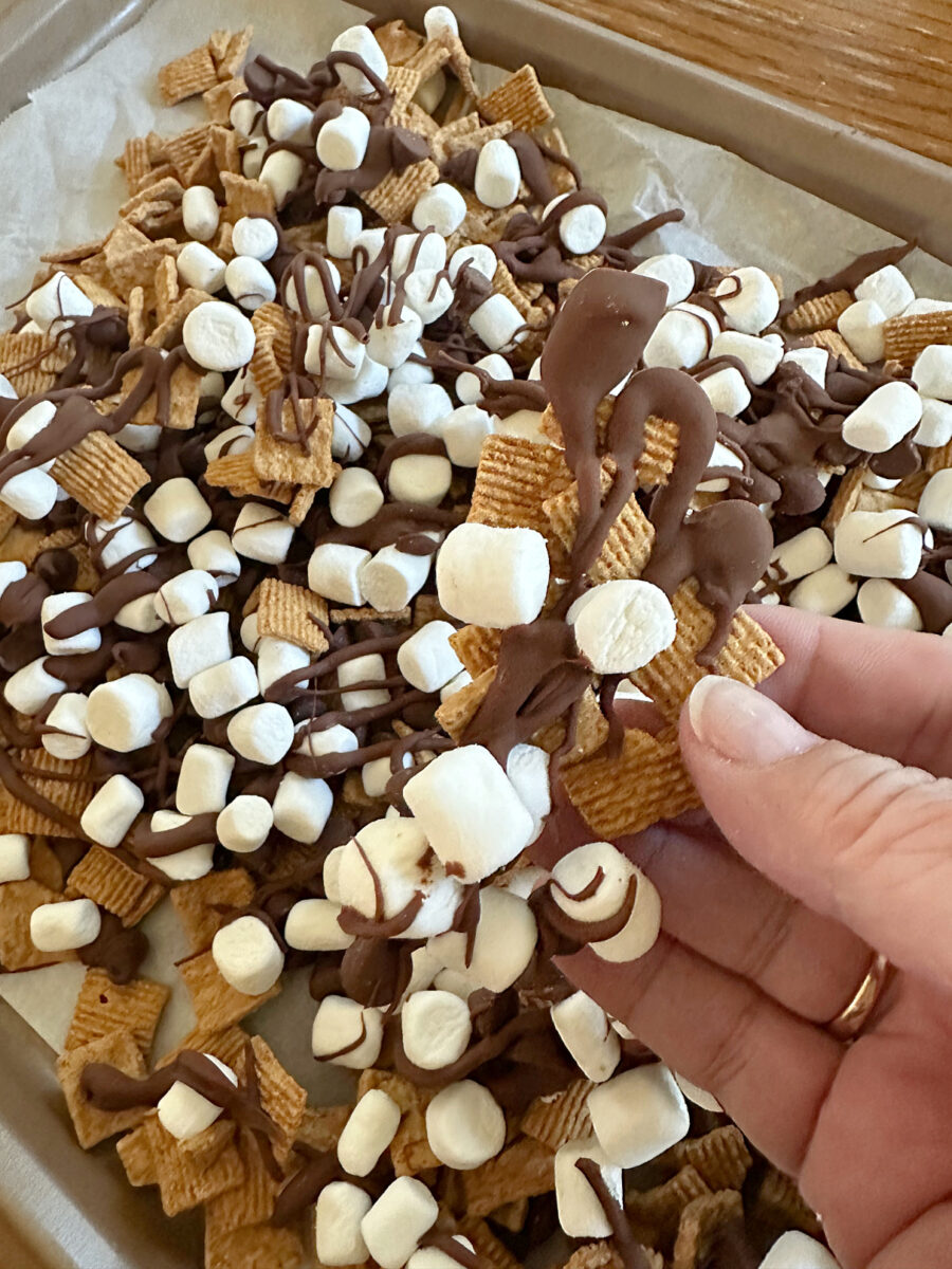 A hand holds a piece of S'mores Snack Mix covered in melted milk chocolate and mini marshmallows above a baking sheet filled with the finished snack. 