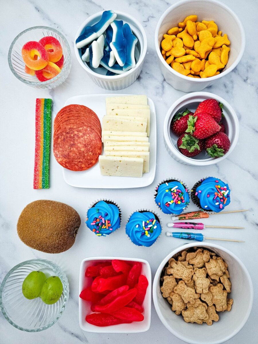 Ingredients needed to make a Pool Party Charcuterie Board including fruit, candy, cheese, pepperoni, cupcakes, Goldfish crackers, and Teddy Grahams.