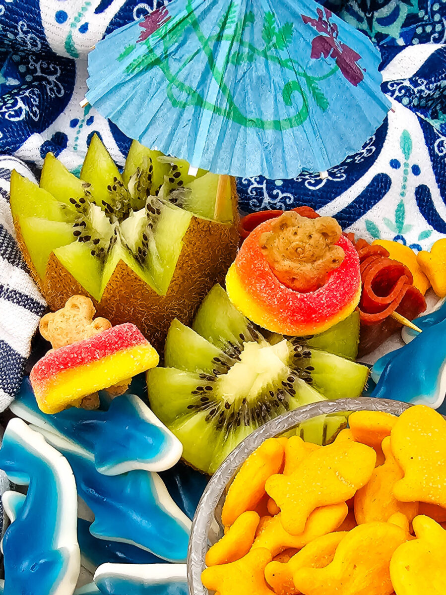 Kiwi fruit cut into flower shapes surrounded by gummy sharks, strawberries, pepperoni, and goldfish crackers on a summer snack board.