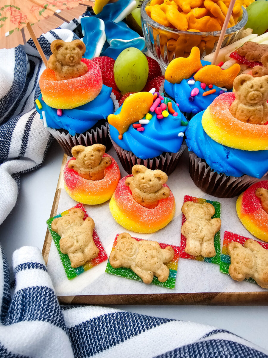 Close-up of teddy graham crackers, gummy candy, and blue frosted cupcakes on a colorful pool party snack board.