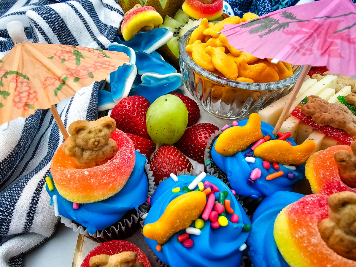 Ocean-inspired summer snack board with blue gummy sharks, fruit, goldfish crackers, cupcakes, and playful pool party decorations.