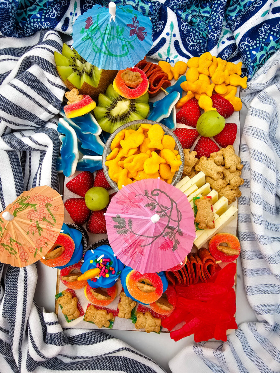 Overhead view of a pool party snack board filled with strawberries, kiwi flowers, cupcakes, cheese, crackers, pepperoni, and colorful summer treats.