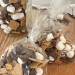 A glass jar and two treat bags are filled with homemade S'mores Snack Mix and displayed on a rustic wood slice. Text overlay reads "S'mores Snack Mix Gift Bags" with the subtitle "Fun Party Favors."