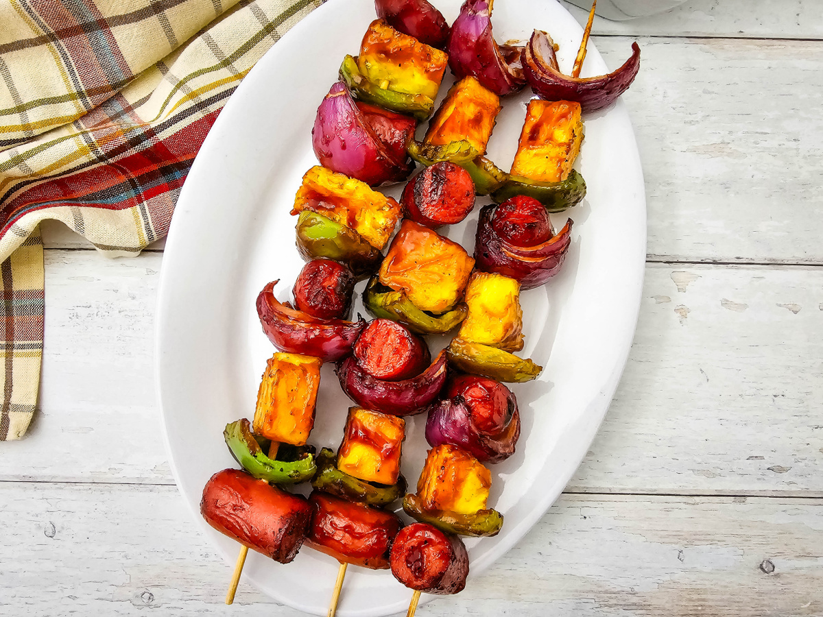 Three grilled hot dog kabobs are served on a white oval platter with caramelized pineapple, grilled peppers, and red onion. A bowl of barbecue sauce and a plaid towel create a backyard barbecue setting.