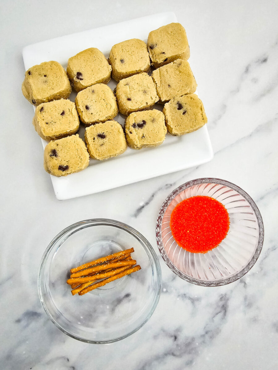 Edible chocolate chip cookie dough pieces, orange sanding sugar, and pretzel sticks arranged for making the no-bake treats.
