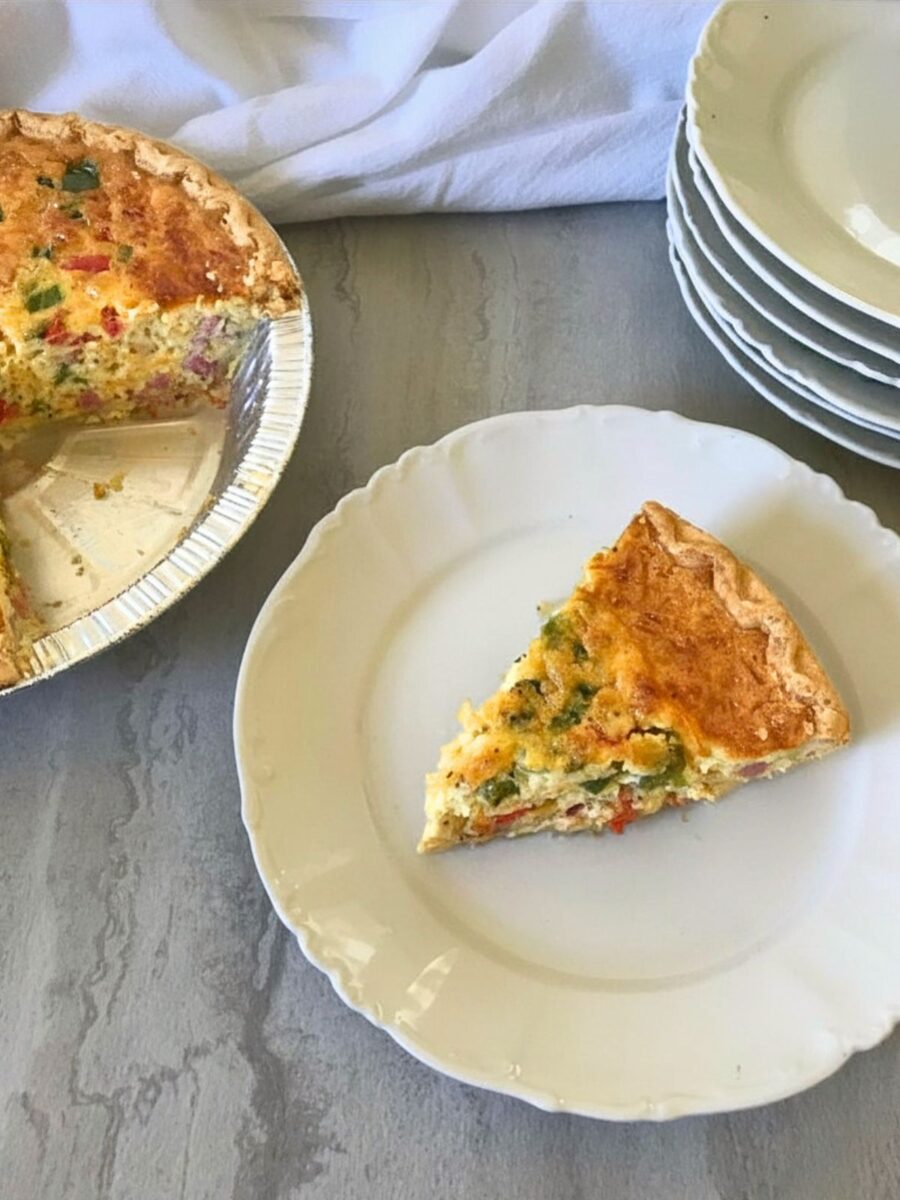 Plated slice of ham and bell pepper quiche beside the remaining breakfast pie and a stack of dishes.
