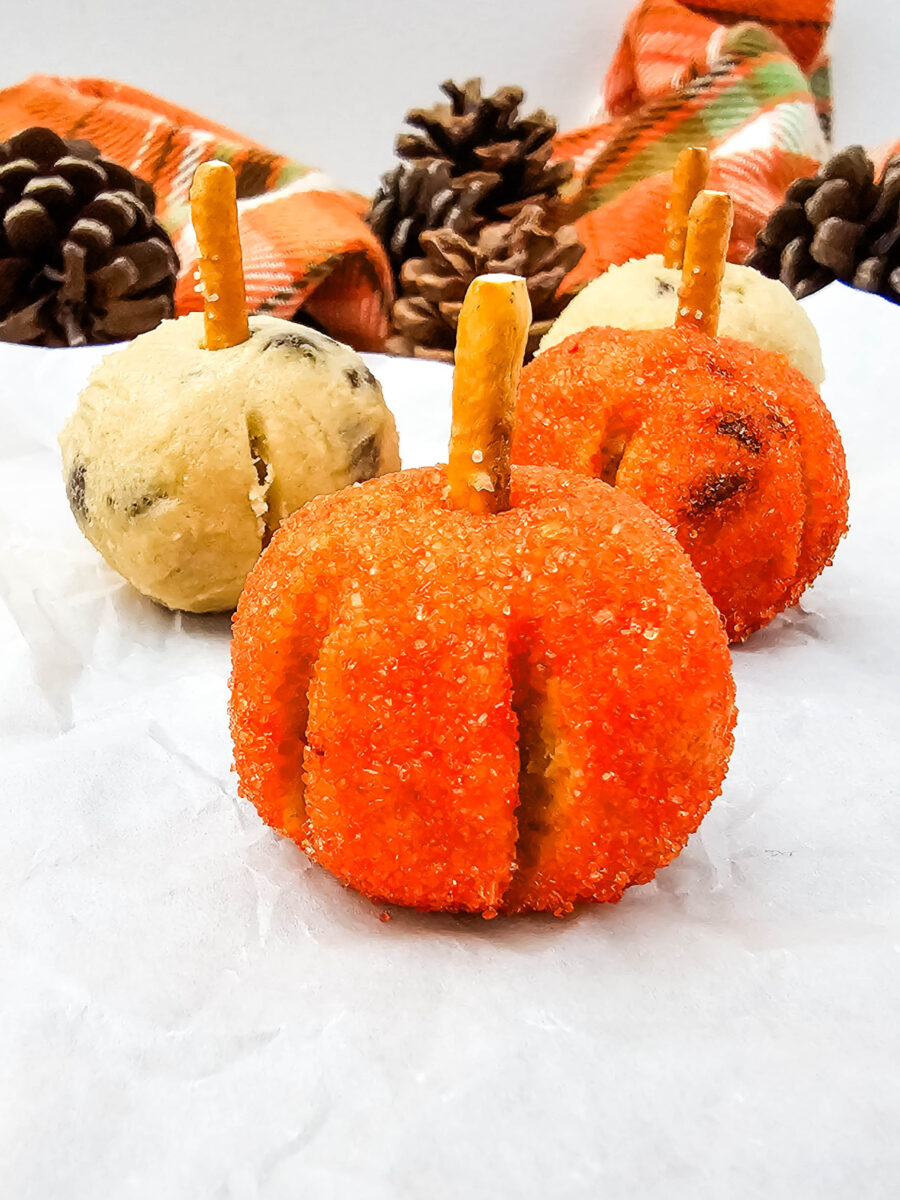 Close-up of orange sugar-coated cookie dough pumpkins with pretzel stems, surrounded by plain cookie dough bites and fall decor.