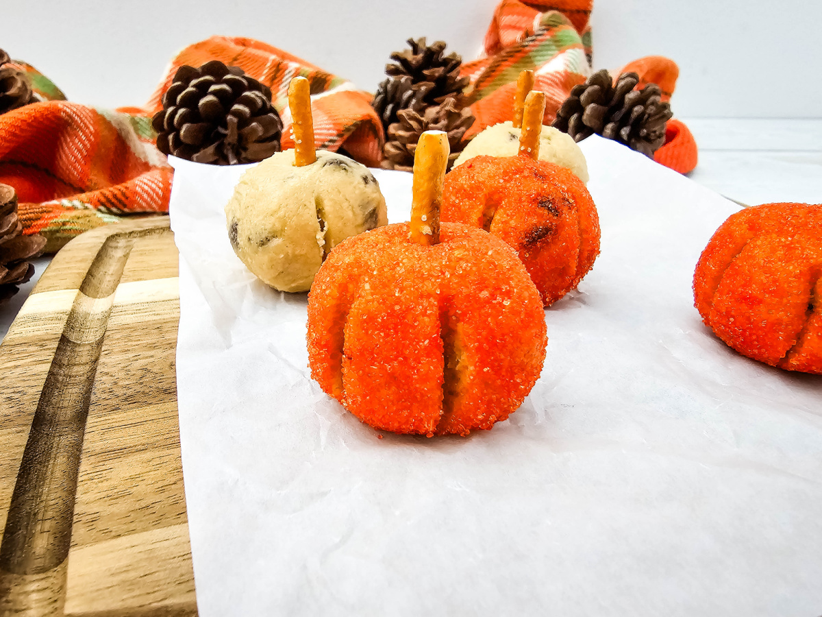 No-bake cookie dough pumpkin balls coated in orange sanding sugar and topped with pretzel stems on parchment paper.