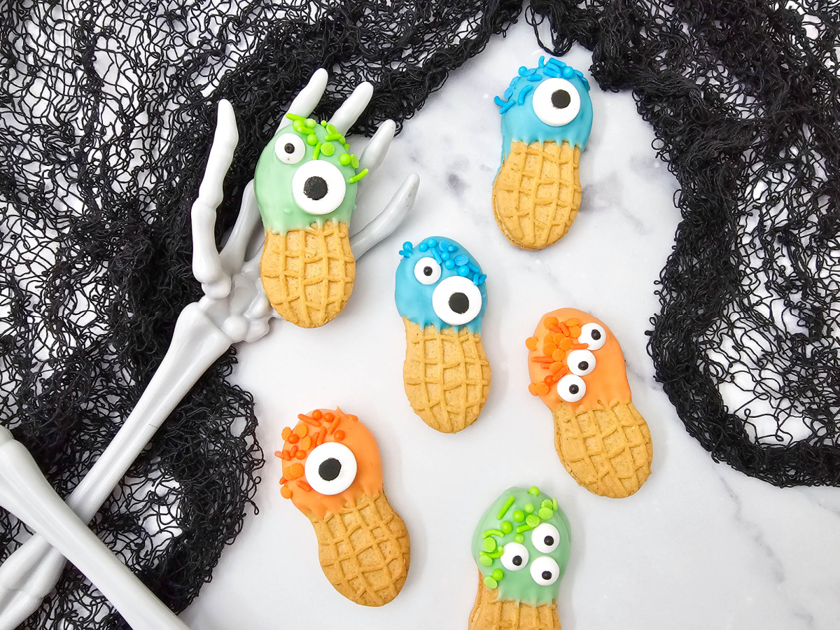 Six Halloween Nutter Butter cookies decorated with colorful candy melts, sprinkles, and assorted candy eyes are arranged between skeleton hands and black lace.