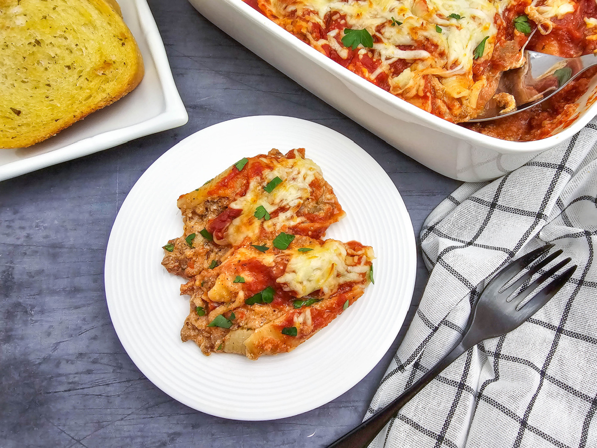Two sausage stuffed shells on a white plate with garlic bread, the casserole dish, a checkered napkin, and black fork in the background.