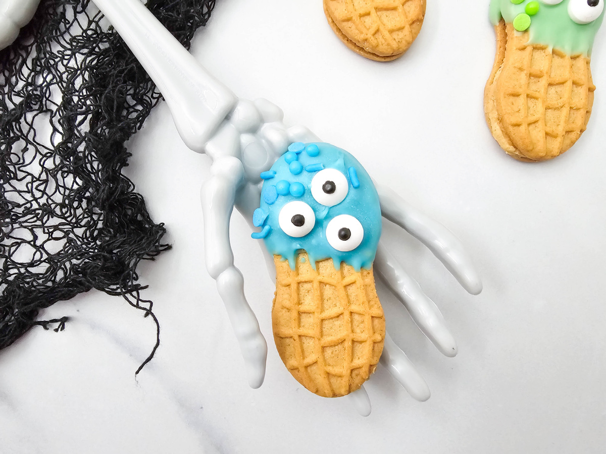 A blue candy-coated Nutter Butter monster cookie with three candy eyes and blue sprinkles rests in a white skeleton hand beside black lace.
