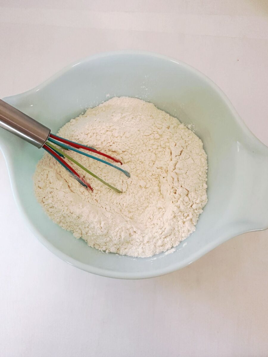 Flour, baking powder, and salt in a light blue mixing bowl with a colorful whisk resting inside.