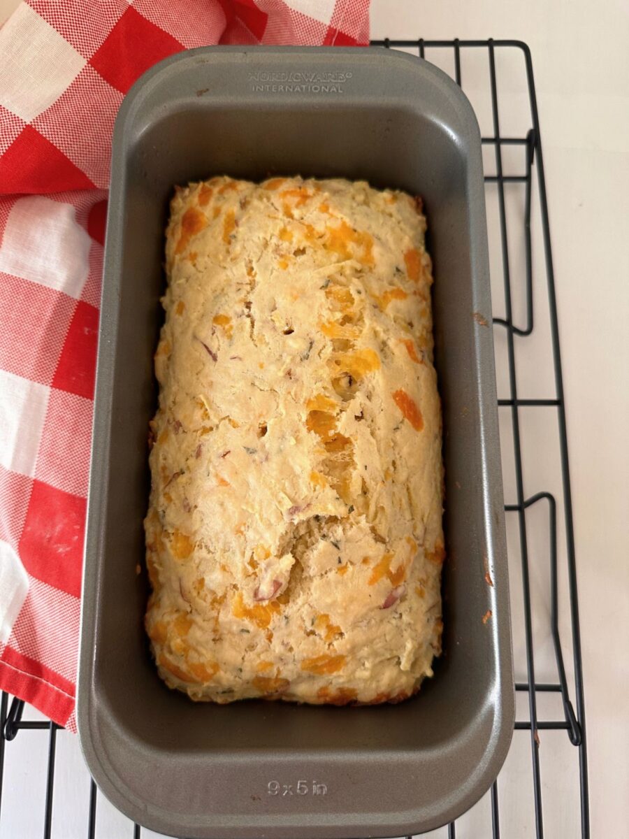Freshly baked apple and cheddar loaf cooling in a gray loaf pan on a black wire rack beside a red-and-white checked towel.
