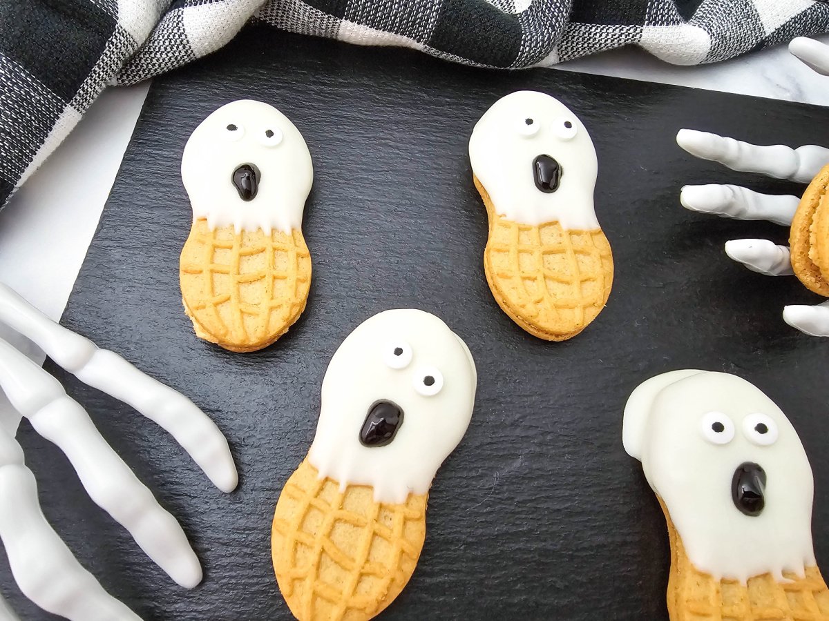 Close-up of four ghost cookies with white candy coating, candy eyes, and different black gel expressions arranged on a black serving tray.