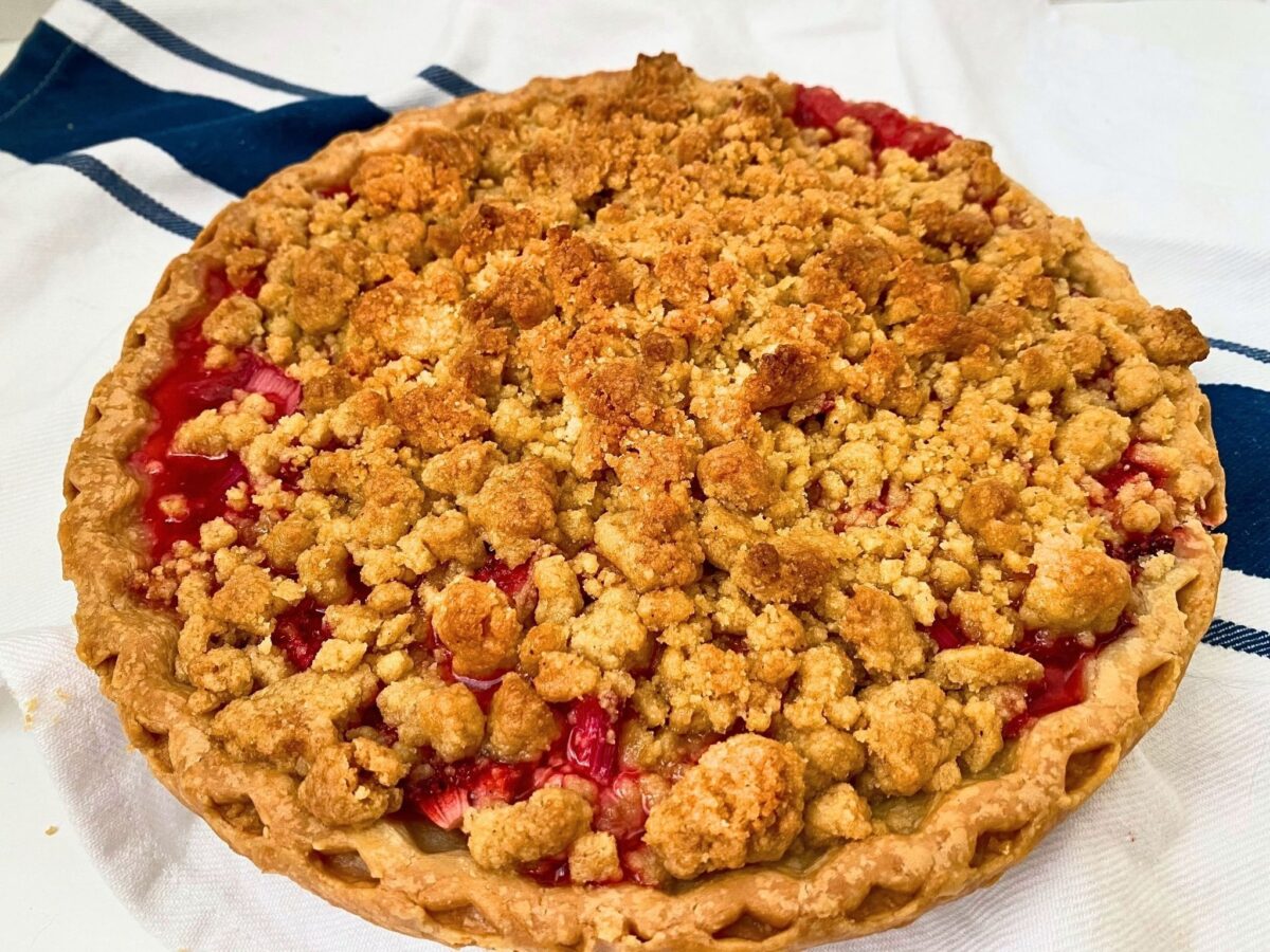 Close-up of a whole strawberry rhubarb pie covered with golden brown sugar crumbs, with the red fruit filling visible around the edges.
