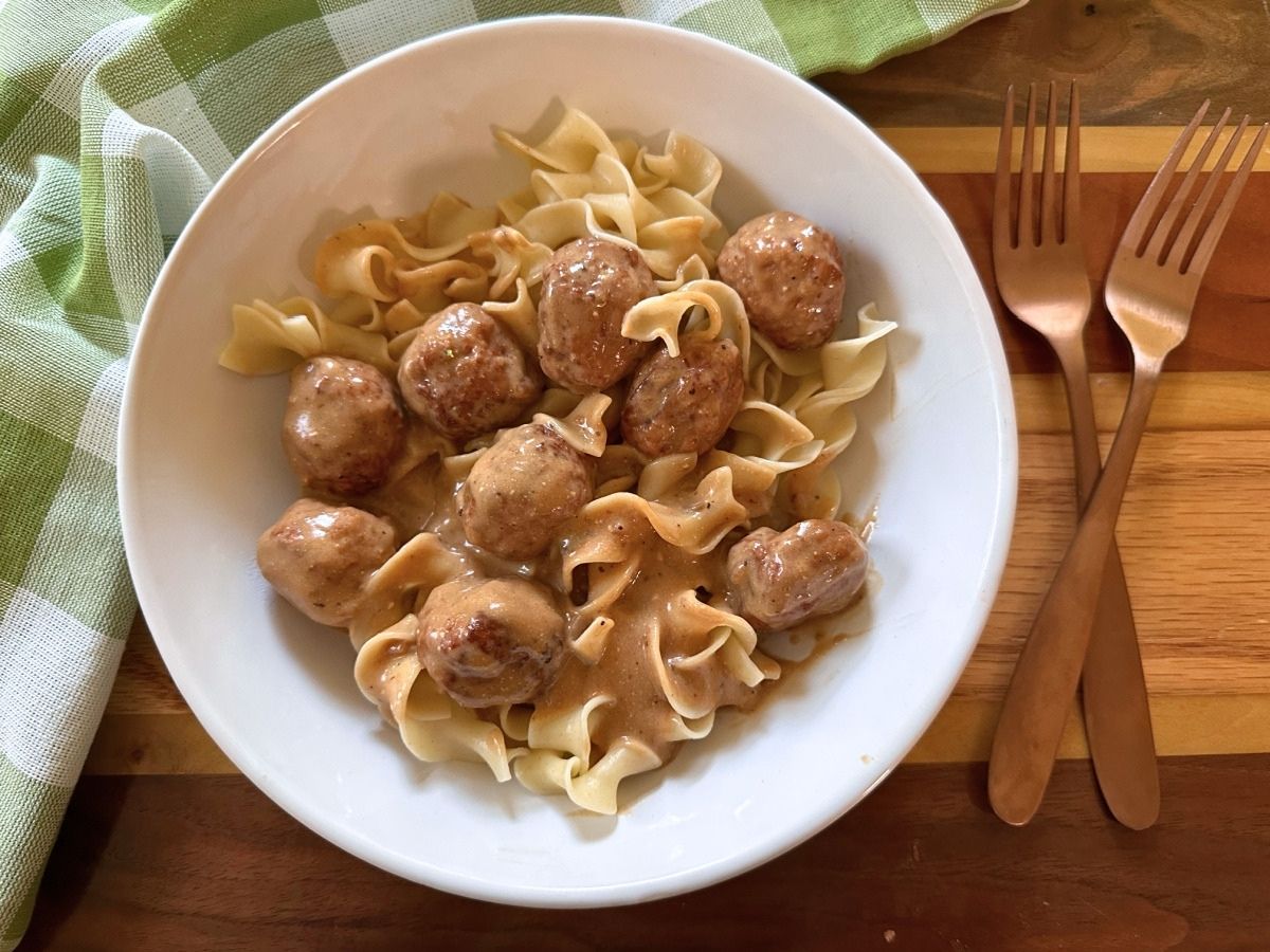 Close-up of Swedish meatballs and egg noodles in a white bowl with creamy sauce spooned over the top.