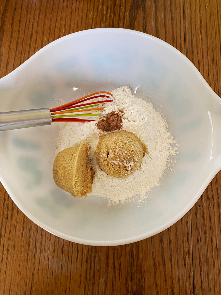 Flour, packed brown sugar, nutmeg, and salt in a white mixing bowl with a whisk, ready to be combined for the crumb topping.
