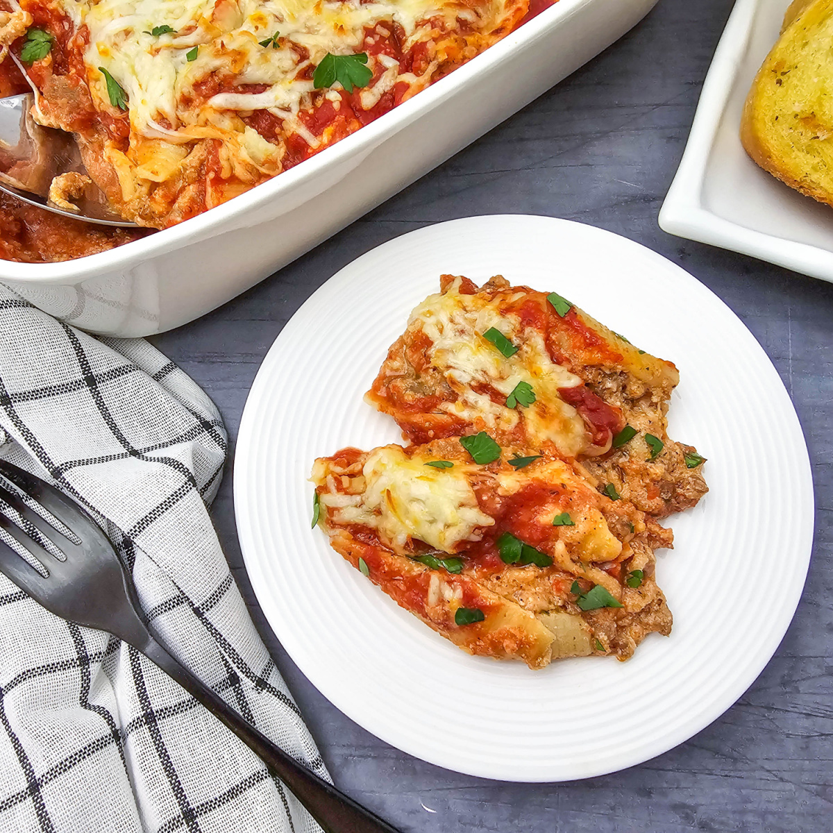 Italian sausage stuffed shells baked with tomato sauce and melted cheese, served on a white plate beside the casserole dish and slices of garlic bread.