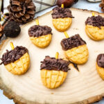 Acorn Nutter Butter Cookies arranged on a rustic wood slice with pinecones, plaid fabric, and decorative acorns.