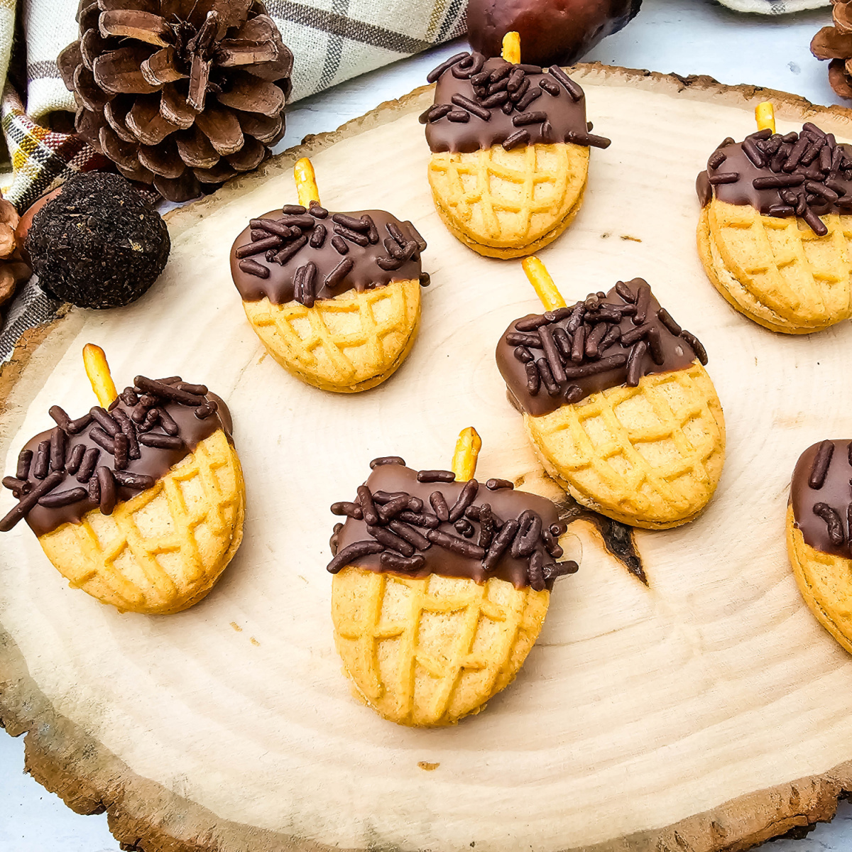 Acorn Nutter Butter Cookies arranged on a rustic wood slice with pinecones, plaid fabric, and decorative acorns.