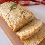Sliced Apple Cheddar Rosemary Bread on a wooden cutting board, garnished with fresh rosemary with apples and a checked cloth in the background.