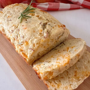 Sliced Apple Cheddar Rosemary Bread on a wooden cutting board, garnished with fresh rosemary with apples and a checked cloth in the background.