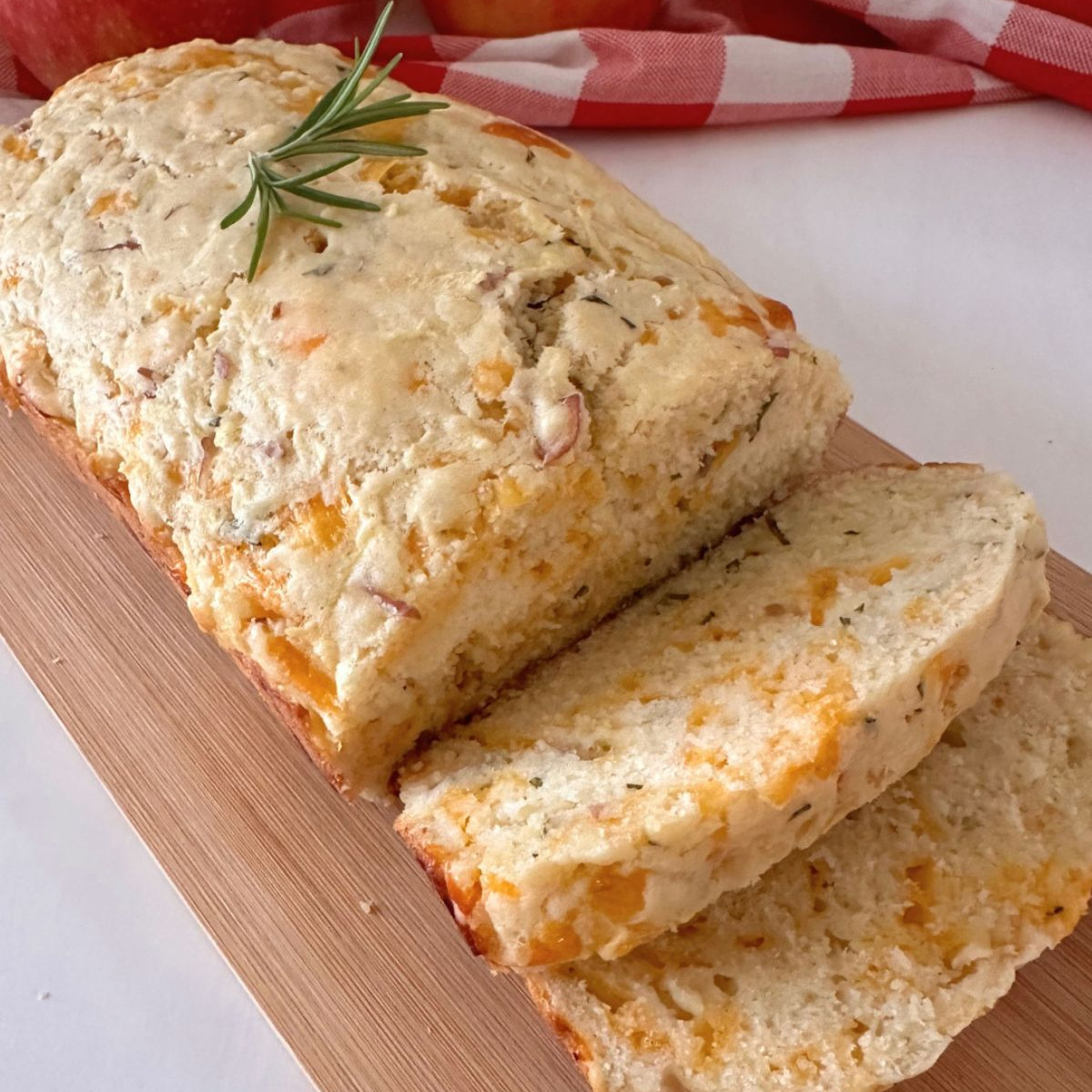 Sliced Apple Cheddar Rosemary Bread on a wooden cutting board, garnished with fresh rosemary with apples and a checked cloth in the background.