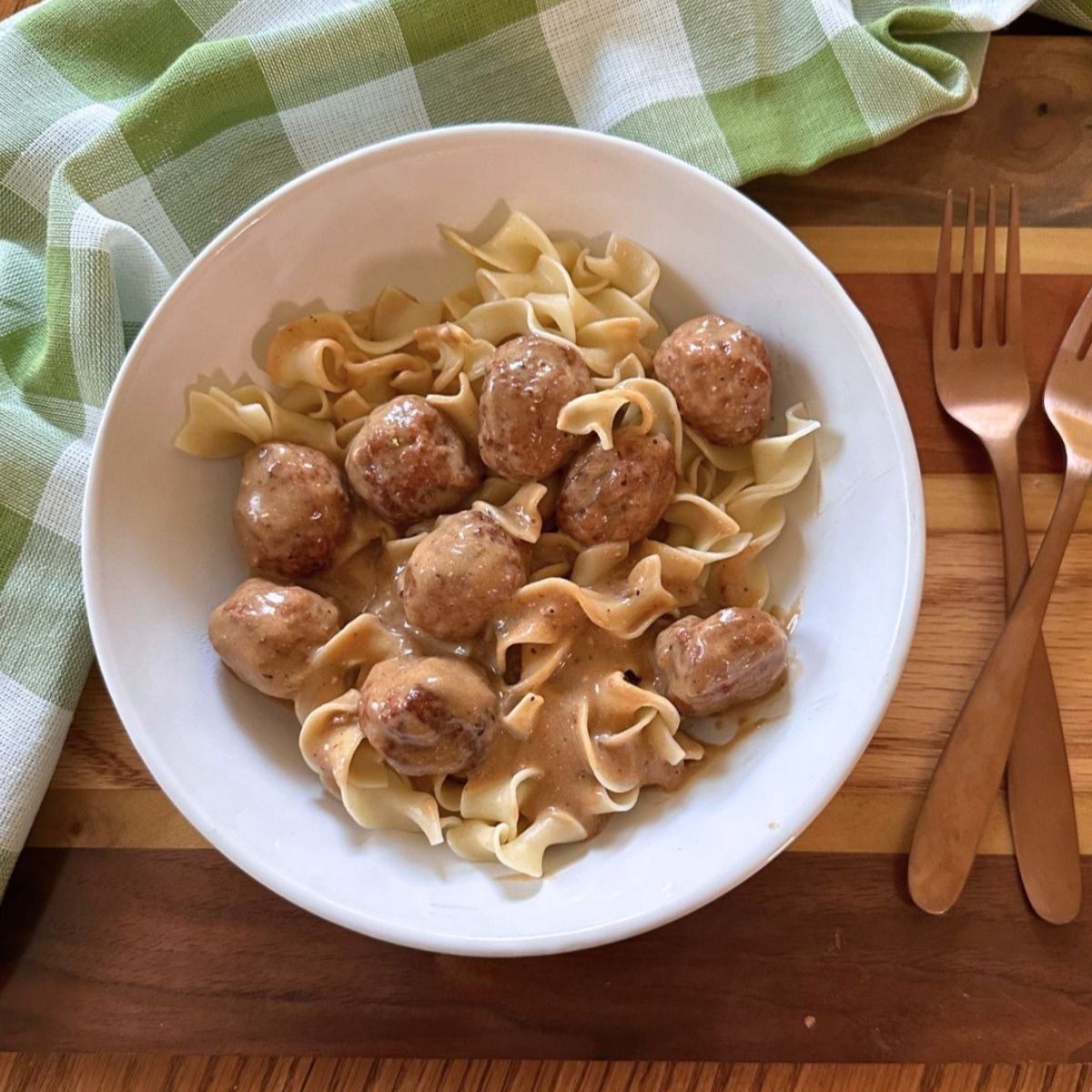Crock Pot Swedish meatballs served over egg noodles in a white bowl beside a green gingham towel and copper forks.