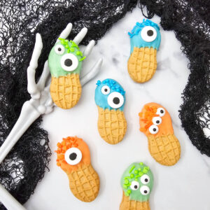 Monster Nutter Butter Cookies decorated with blue, green, and orange candy melts, matching sprinkles, and assorted candy eyes are arranged with skeleton hands and black lace.