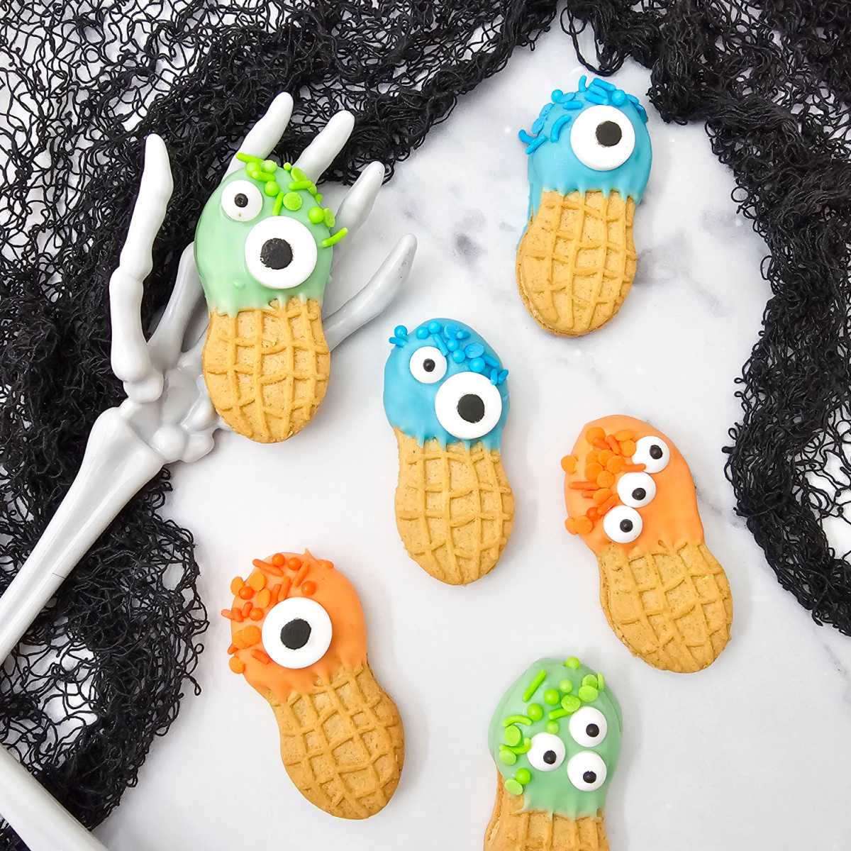 Monster Nutter Butter Cookies decorated with blue, green, and orange candy melts, matching sprinkles, and assorted candy eyes are arranged with skeleton hands and black lace.