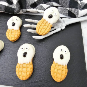 Nutter Butter Ghost Cookies dipped in white candy coating and decorated with candy eyes and black gel mouths on a black slate tray with a skeleton hand.